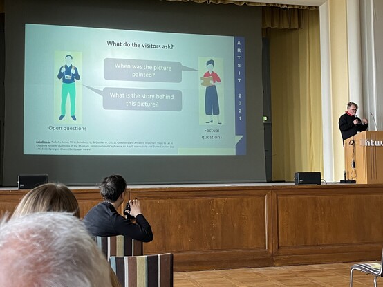 A person giving a presentation in a conference room with a projection screen displaying a slide about visitor questions in a museum context, with attendees watching and one person taking a photograph.