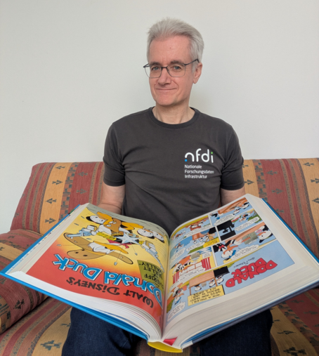 Cord Wiljes, reading the book "Disney Comics Library. Carl Barks’ Donald Duck. Vol. 1. 1942–1950" by Carl Barks, pblished by Taschen, sitting on a sofa, wearing an NFDI t-shirt.