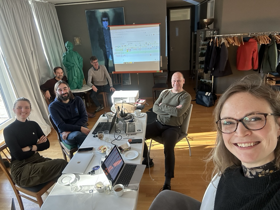 Das Team der Cultural Research Data Academy sitzt in einem Seminarraum in Eltville vor der einer Leinwand. In der Mitte befindet sich ein Tisch mit Laptops. Im Hintergrund ist eine Figur von Goethe zu sehen.