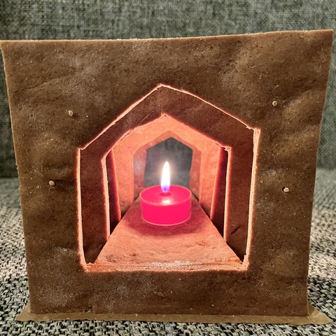 Ein Lebkuchenhaus mit einer Reihe von Bögen, die einen geschichteten Effekt erzeugen. Eine kleine rote Kerze leuchtet im Inneren und wirft ein warmes Leuchten durch die Öffnungen.