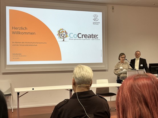 Eine Konferenz mit zwei Personen auf einem Podium. Eine Rednerin präsentiert, während der andere neben ihr steht. Auf einem großen Bildschirm wird der Titel "CoCreate: Wissen gemeinsam organisieren - Open Science" zusammen mit einer Begrüßungsnachricht angezeigt.