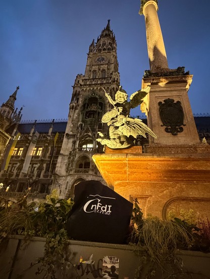 Die NFDI4Culture-Tasche liegt am beleuchteten Brunnen vor dem Neuen Rathaus in München vor einem blauen Abendhimmel.
