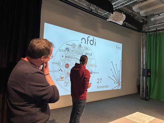 Kai Matuszkiewicz (FID Media) hört sich die Erläuterungen von Alexander Stark (NFDI4Culture) zum Netzwerk der NFDI-Konsortien an.