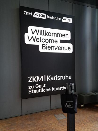 Foto von Displayanzeige "ZKM Karlsruhe, Willkommen, Welcome, Bienvenue, ZKM, Karlsruhe zu Gast Staatliche Kunsthalle". Vorne wurde an einer kurzen Säule die NFDI4Culture-Tasche aufgehängt. Fotos by Katja Sternitzke, CC-0