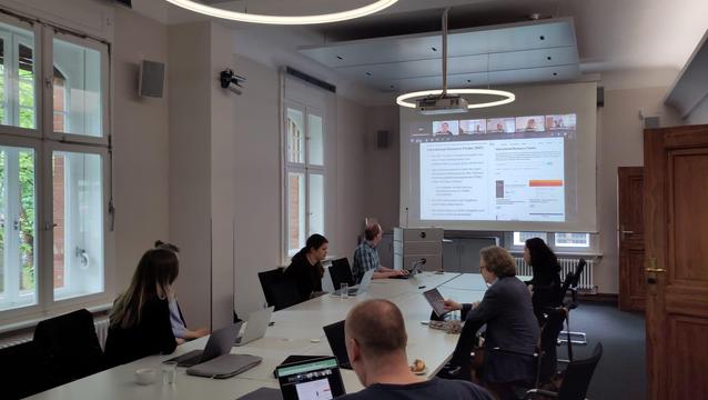 Die Teilnehmer:innen des Treffens der DHd-AG Referenzcurriculuum sitzen an einer langen Tischreihe und schauen gemeinsam auf eine Leinwand, wo eine Präsentation projiziert ist. 