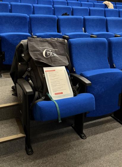Reihen von blauen Kinosesseln, auf einem Randplatz ist ein Rucksack mit dem NFDI4-Culture-Beutel platziert, davor liegt ein Blatt Papier mit dem Workshop-Programm. Photo Credit: Martin Albrecht-Hohmaier